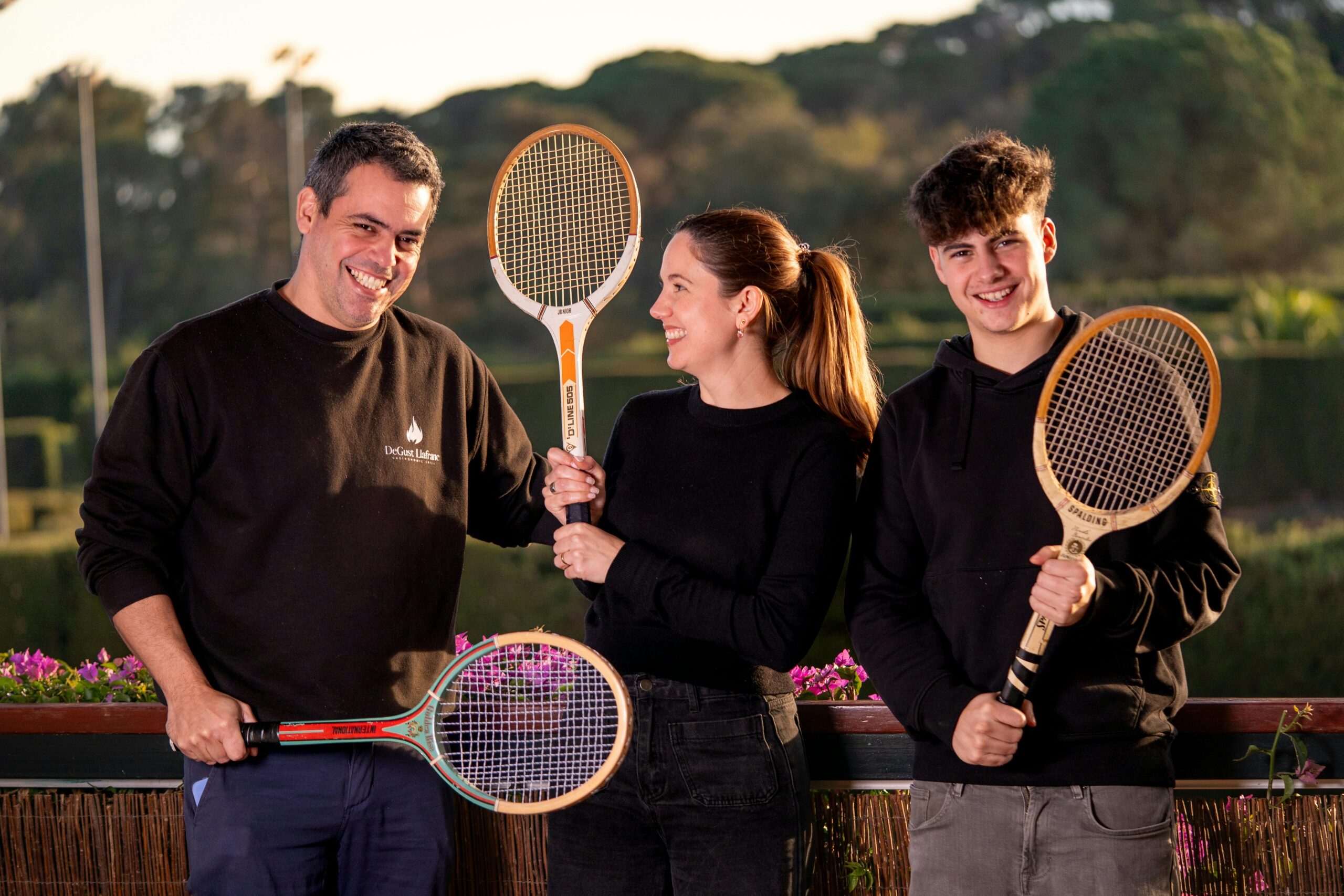 Drei lÃ¤chelnde Personen halten alte TennisschlÃ¤ger vor einem AuÃŸenhintergrund.