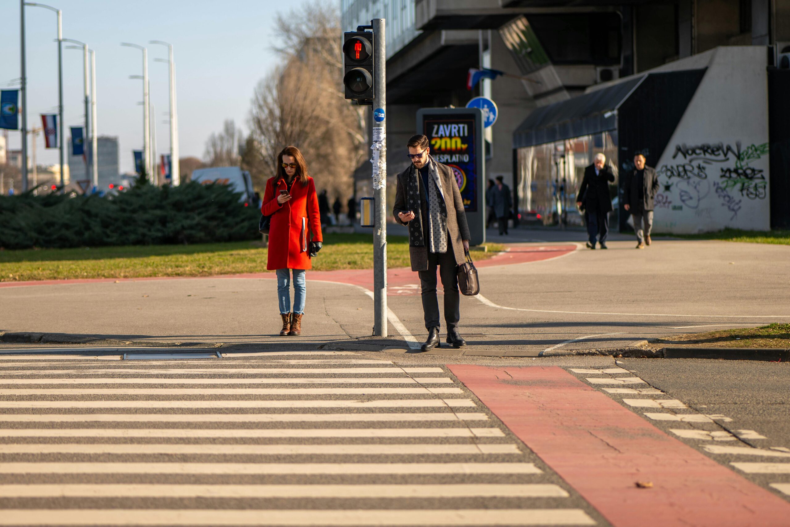 Zwei Erwachsene Ã¼berqueren eine StraÃŸe in Zagreb, aufgenommen bei Tageslicht an einem klaren Tag.